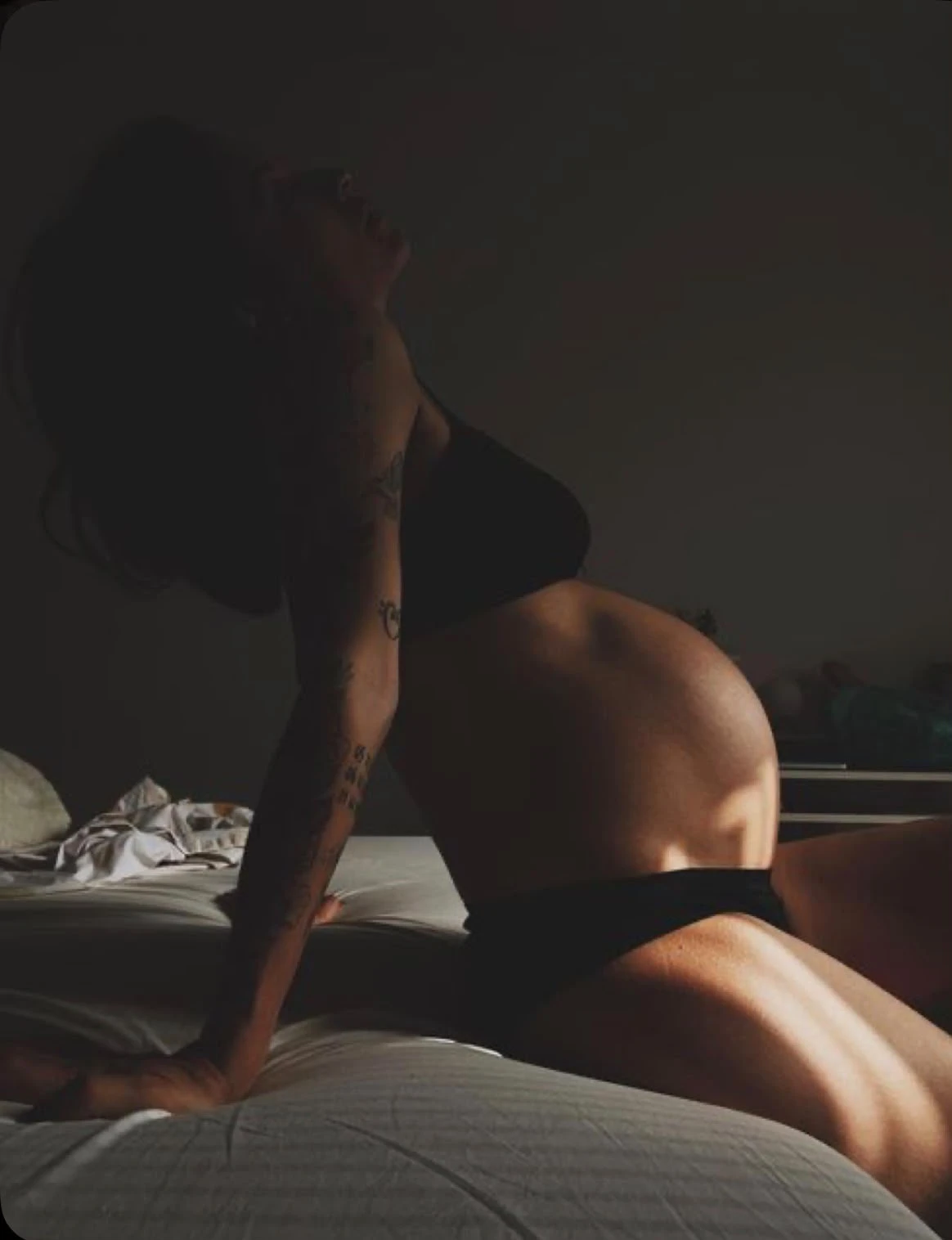 A heavily pregnant woman kneeling on a bed in low light, head tipped back